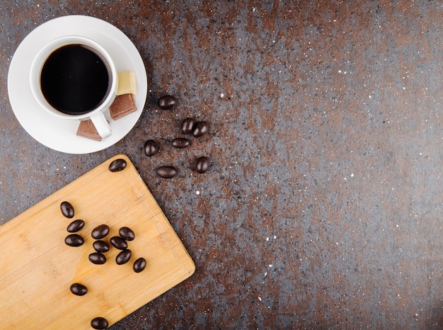 Top view of glazed chocolate nut candy scattered on a wooden board and a cup of coffee on black background with copy space