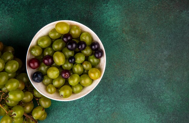 Top view of grape berries in bowl and bunch of grape on green background with copy space