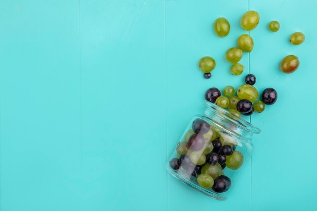 Top view of grape berries spilling out of glass jar on blue background with copy space