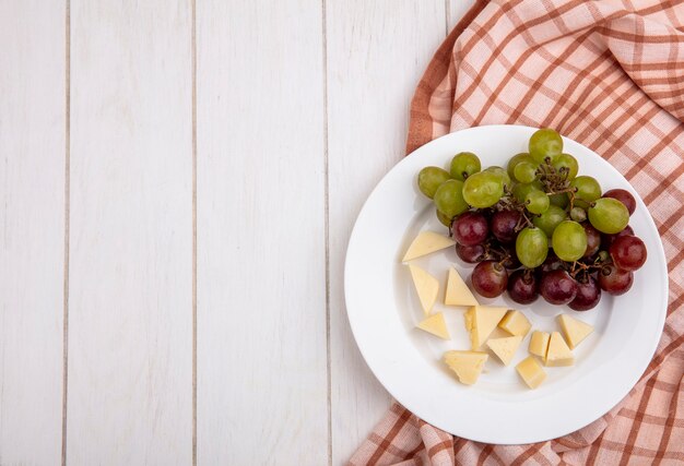 Вид сверху на виноград и нарезанный сыр в тарелке на клетчатой ткани на деревянном фоне с копией пространства