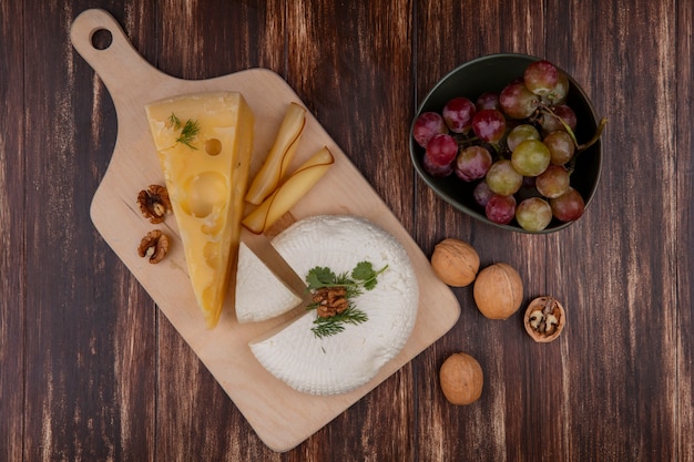 Top view grapes in a bowl with maasdam and feta cheese and nuts on a stand against a wooden background