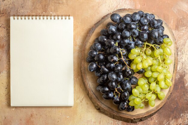 Вид сверху на виноградную деревянную доску с зеленым и черным виноградом рядом с белой записной книжкой