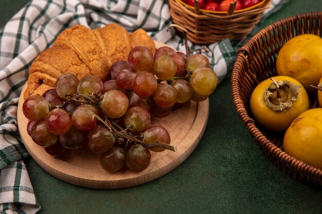 Top view of grapes on a wooden kitchen board on a checked cloth with croissant with persimmon fruits on a bucket on a green background