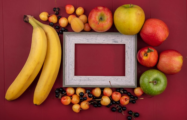 Free photo top view of gray frame with bananas black currants apples peaches and white cherries on a red surface