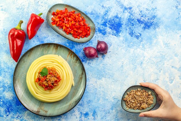 Top view of a gray plate with delicious spagetti chopped and whole red pepper onions and hand holding meat on blue table