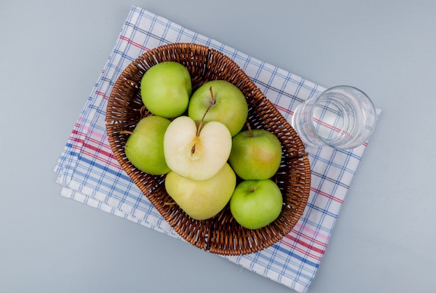 Вид сверху зеленых яблок в корзине и стакане воды на клетчатой ткани и сером фоне