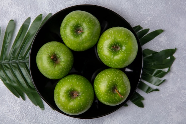 Вид сверху зеленые яблоки на черной тарелке на ветвях с листьями на белом фоне