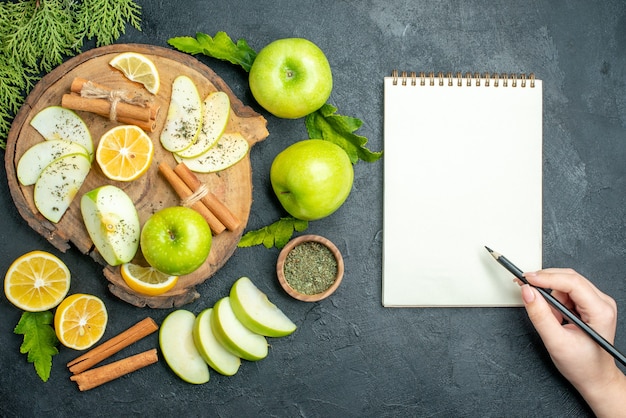 위쪽 보기 녹색 사과 계피 스틱과 레몬 조각 나무 판자에 있는 사과 조각은 검은 탁자에 있는 여성 손에 레몬 사과 공책 연필을 자른다