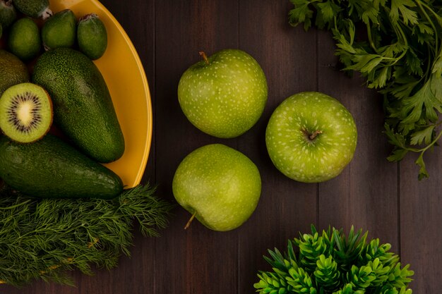 Top view of green apples with fresh fruits such as avocados feijoas and kiwis on a yellow plate on a wooden wall