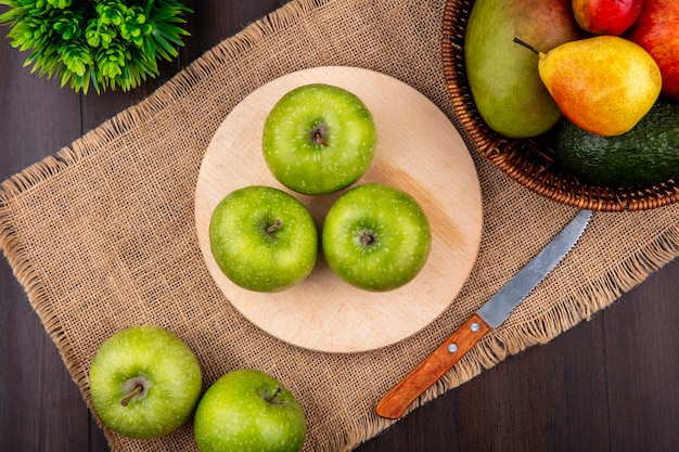 Free photo top view of green apples on a wooden kitchen board with knife on sack cloth with a bucket of fruits on wooden surface