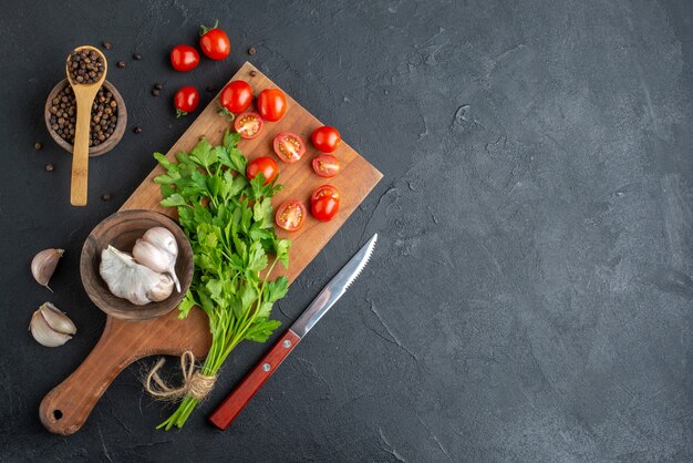 Top view of green bundle fresh whole cut tomatoes garlics on wooden cutting board knife pepper on black distressed surface