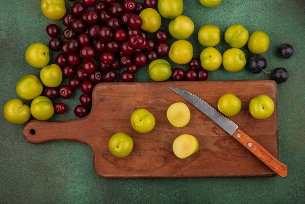 Top view of green cherry plum on a wooden kitchen board with knife with red cherries on a green background