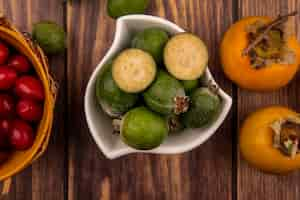 Free photo top view of green feijoas on a bowl with cornelian cherries on a bucket with persimmon fruits isolated on a wooden wall