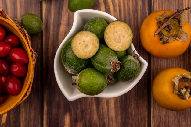 Top view of green feijoas on a bowl with cornelian cherries on a bucket with persimmon fruits isolated on a wooden wall