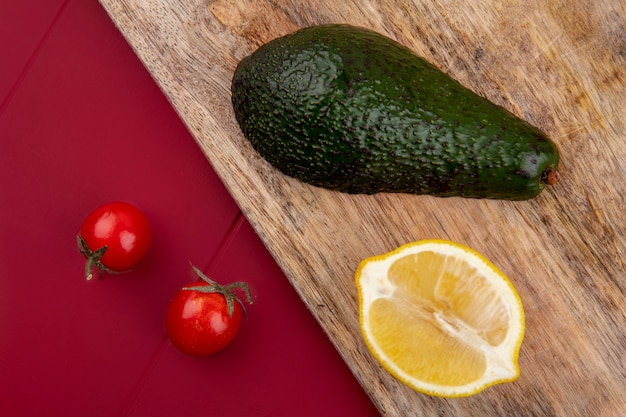 Вид сверху зеленого и свежего авокадо на деревянной кухонной доске с ломтиком лимона и помидорами черри на красной поверхности