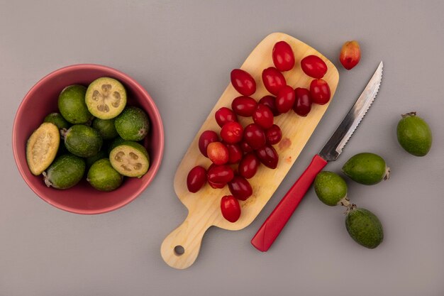 Top view of green fresh feijoas on a bowl with cornelian cherries on a wooden kitchen board with knife on a grey wall