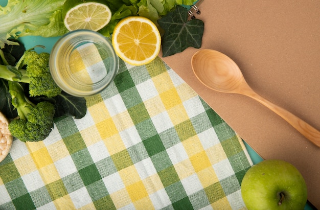 Vista dall'alto verde frutta e verdura brocoli lattuga edera foglie bicchiere d'acqua cucchiaio di legno fetta di mela di limone e lime con copia spazio sulla tovaglia