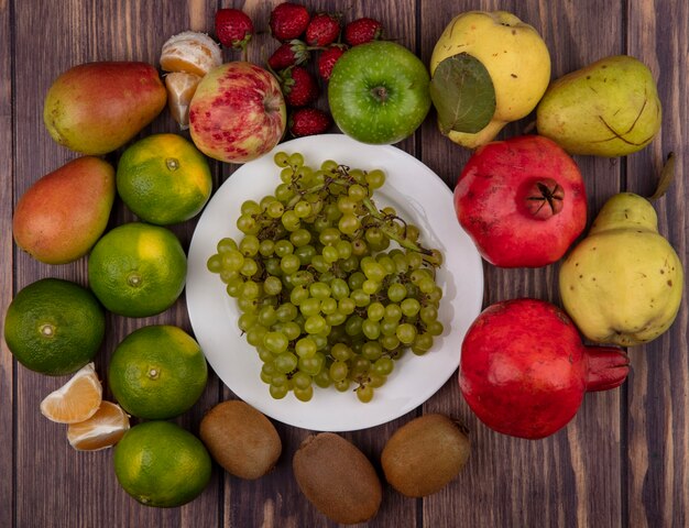 Top view green grapes on a plate with kiwi tangerines pears strawberries and pomegranates on a wooden wall