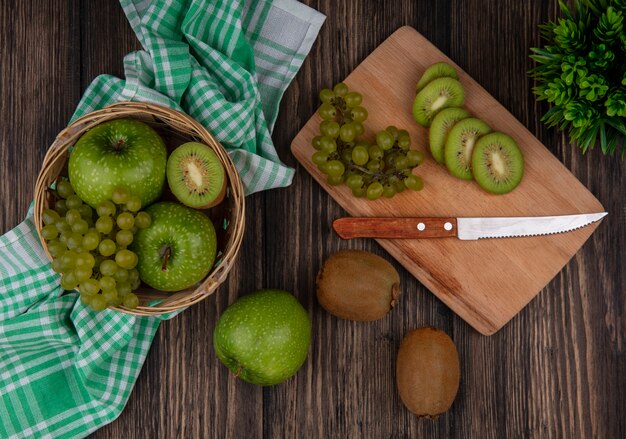 Top view green grapes with kiwi slices and a knife on a board and green apples in a basket on a green checkered towel on a wooden background