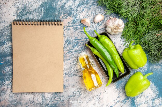 Top view green hot peppers on black plate garlic peppers bottle of oil a notebook on blue-white background