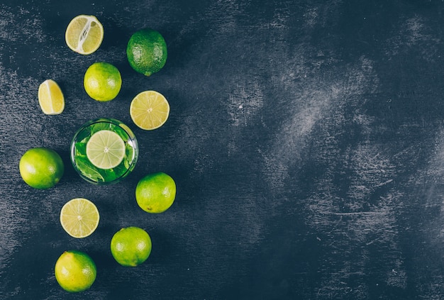 Free photo top view green lemons in water glass and around with slices on black textured background. horizontal space for text