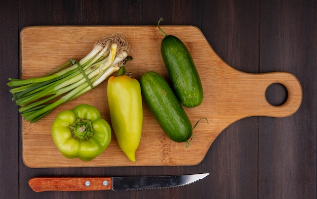 Foto gratuita vista dall'alto cipolle verdi con cetrioli e peperoni verdi su un tagliere con un coltello su un fondo di legno
