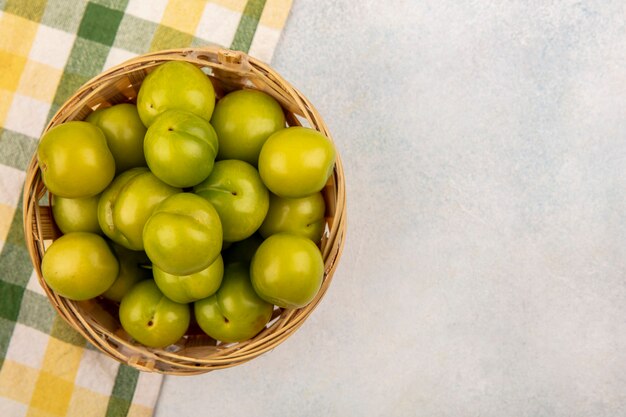 Top view of green plums in basket on plaid cloth on white background with copy space