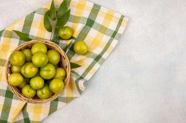 Top view of green plums in basket with leaves on plaid cloth and white background with copy space