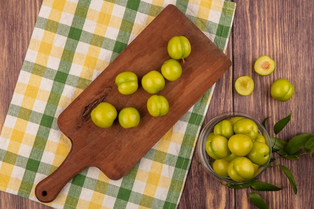 Top view of green plums on cutting board on plaid cloth and in jar on wooden background