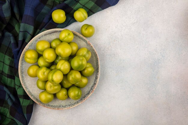 Top view of green plums in plate on plaid cloth and on white background with copy space