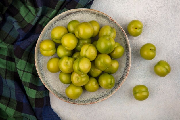 Top view of green plums in plate on plaid cloth and on white background