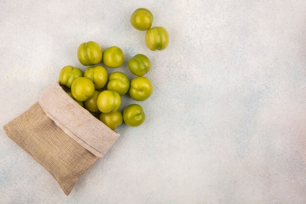 Top view of green plums spilling out of sack on white background with copy space