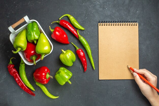Top view green and red peppers in plastic basket hot peppers a notebook pencil in woman hand on dark surface