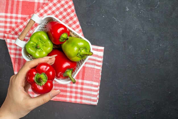 Top view green and red peppers in plastic basket on red white checkered tablecloth red bell pepper in female hand on dark surface free space
