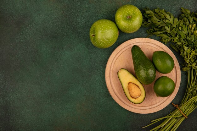 Top view of green skinned avocados on a wooden kitchen board with limes with green apples and parsley isolated on a green surface with copy space