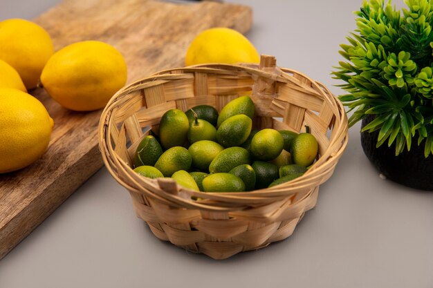 Top view of green skinned kinkans on a bucket with lemons on a wooden kitchen board on a grey background