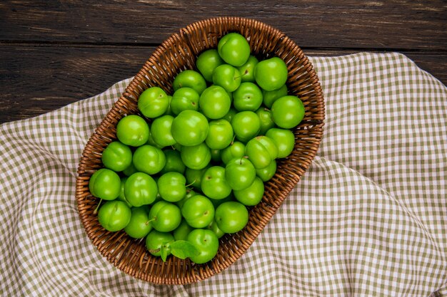 Top view of green sour plums in a wicker basket on plaid fabric on dark wooden table