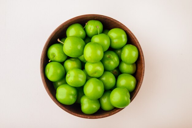 Top view of green sour plums in a wooden bowl on white table