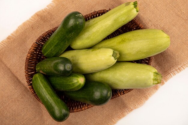 Top view of green vegetables such as cucumbers zucchinis on a bucket on a sack cloth on a white wall