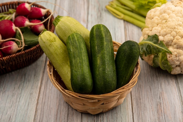 Top view of green vegetables such as cucumbers and zucchinis on a bucket with radishes on a bucket with cauliflower and celery isolated on a grey wooden wall