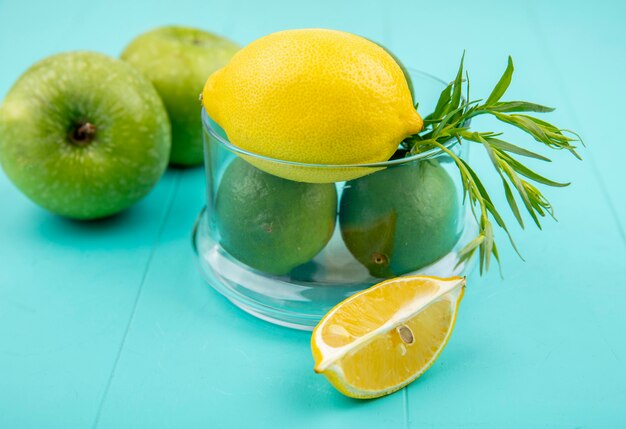 Top view of green and yellow lemons on a glass bowl with green apple on blue surface