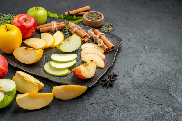 Top view of green yellow and red sliced and whole fresh apples on a black tray and cinnamon limes on the right side on a dark background