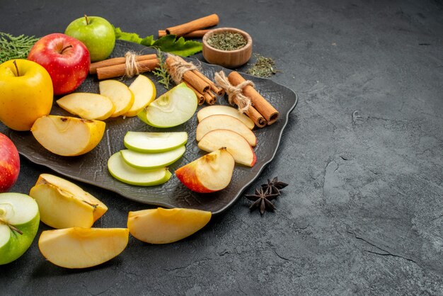 Top view of green yellow and red sliced and whole fresh apples on a black tray and cinnamon limes on the right side on a dark background