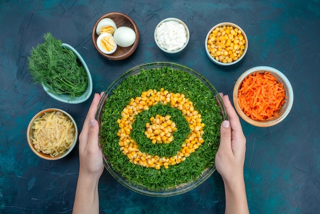 Top view greens salad with corns inside round glass plate with eggs and sliced carrots on the dark blue desk.