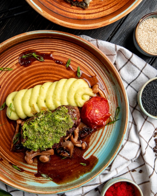 Top view of grilled beef with mashed potatoes tomato mushrooms and avocado sauce in a plate on wood