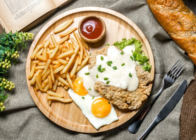 Top view grilled chicken fillet with sauce fried egg ketchup spring onion lettuce and french fries on a board