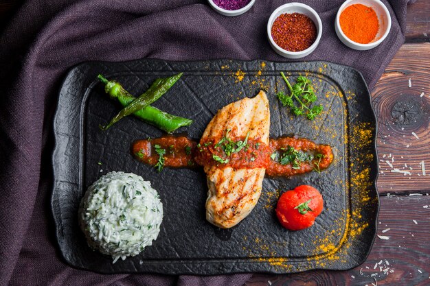 Top view grilled chicken steak with garnish, tomato, pepper a dark wooden table