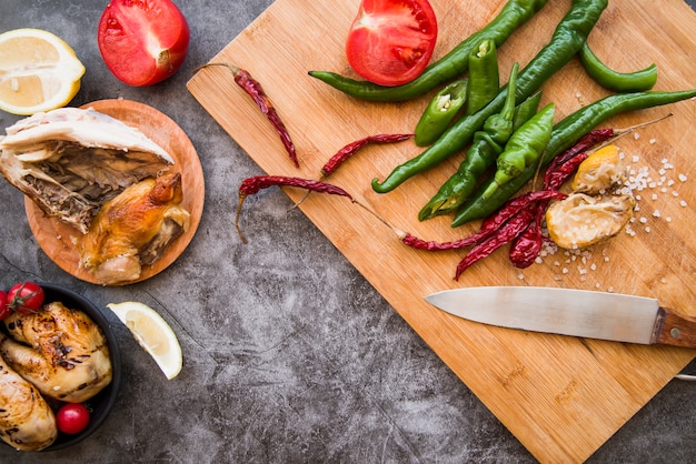 Top view of grilled chicken with green and red chilies on wooden chopping board with knife