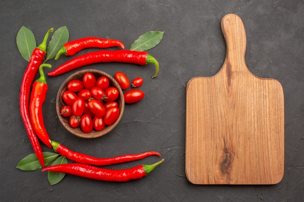 Top view half circle of red hot peppers and bay leaves and a bowl of cherry tomatoes and a chopping board on the black table