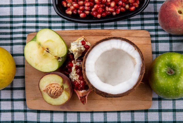 Top view of half coconut on a wooden kitchen board with half apple half peach and pomegranate on checked tablecloth surface